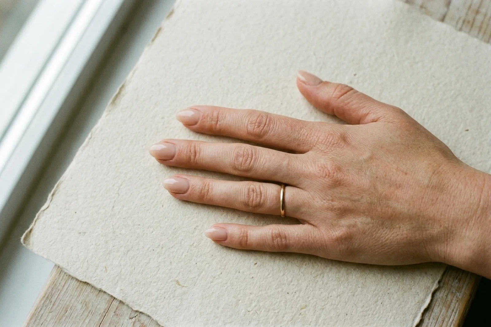 Hånd sett ovenfra på et ark håndlaget papir, med korte avrundede negler i en nude tone der den frie kanten så vidt går utenfor fingertuppen